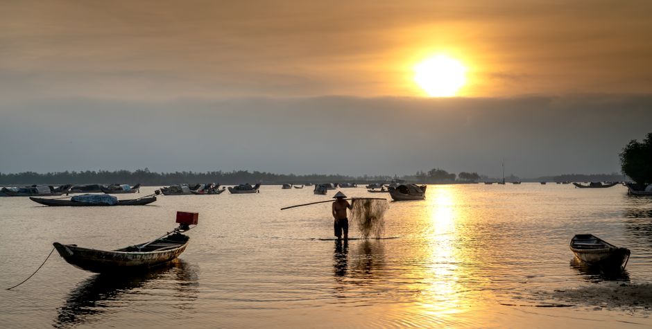 A lone fly fisherman casts a line from a skiff in a serene, golden-lit salt marsh, surrounded by tall spartina grass.